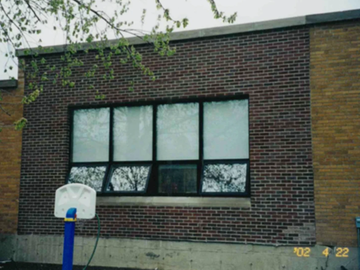 A section of dark red brown brick around a wide window bay in April 2002, meeting a wall of tan brick at a vertical joint on the right, with a child-height basketball post on the paved yard in front