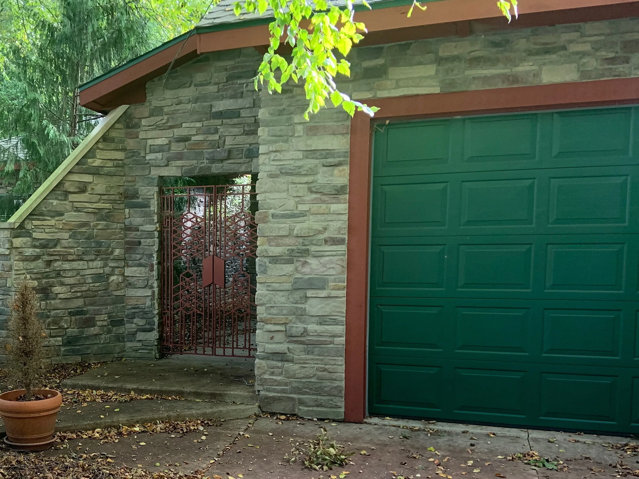 The gated stone entry and green garage door of the home in 2019, burgundy units scattered through grey and tan coursed stone under a red brown fascia, a potted tree on the step