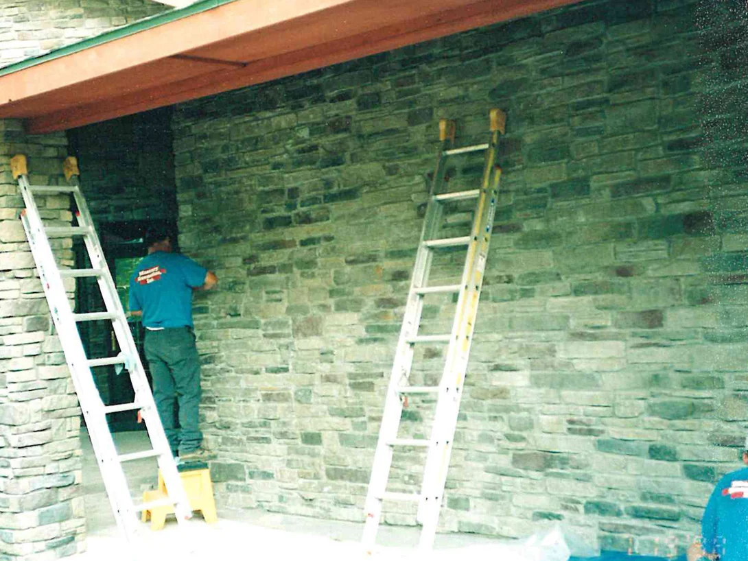 An applicator on a ladder in 1998 working stain into the grey and tan stone wall beside the entry door, a second ladder leaning on the wall and a tarp on the ground