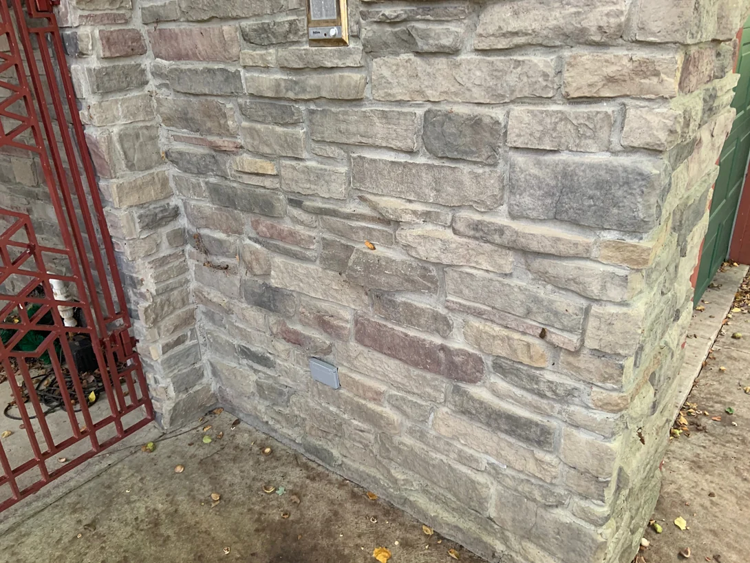 The stone wall beside the entry gate in 2019 up close, burgundy units sitting among grey and tan stone with an intercom and an outlet cover on the wall