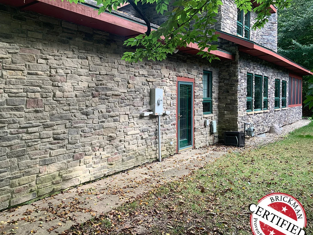The long side elevation of the home in 2019, coursed stone running past a glazed door and a row of windows under a red brown eave, with the certification stamp in the corner