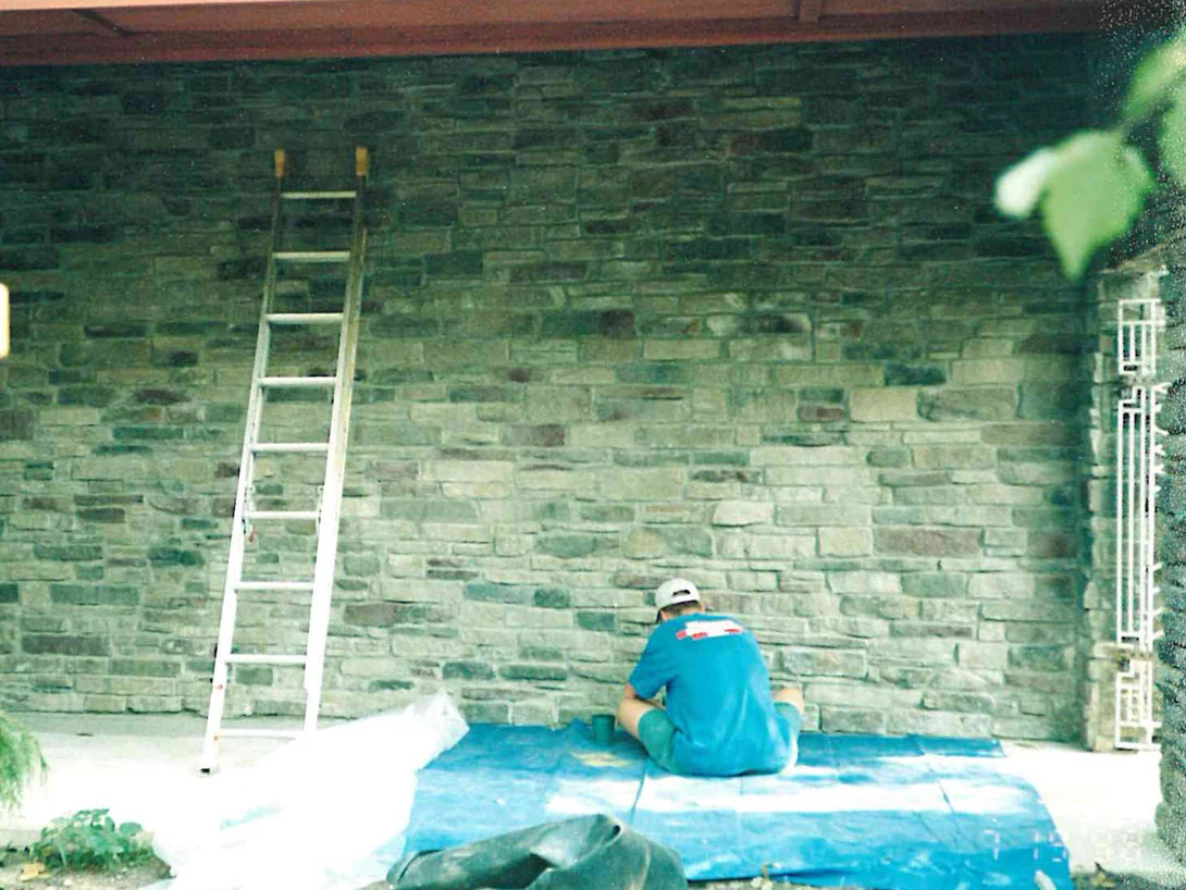 An applicator seated on a blue tarp in 1998 staining the base of the stone wall under the porch roof, a ladder leaning on the wall beside him
