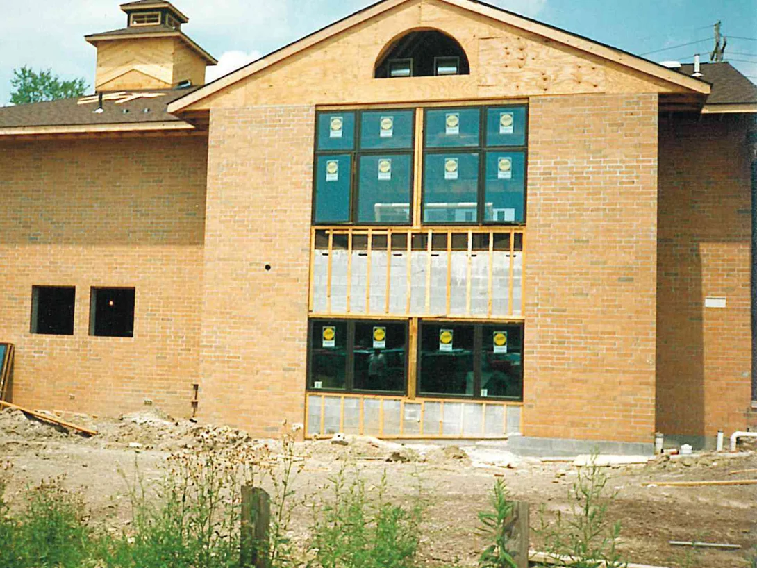 The new addition in 1996, its gable end sheathed in raw plywood above unglazed openings, the brick below a pale orange tan