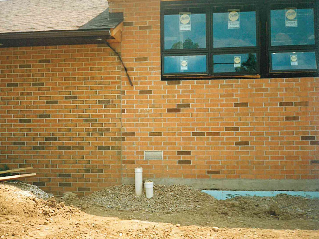 A low wall of the addition in 1996 with darker units set through a pale orange tan field, bare earth and pipe stubs at its base