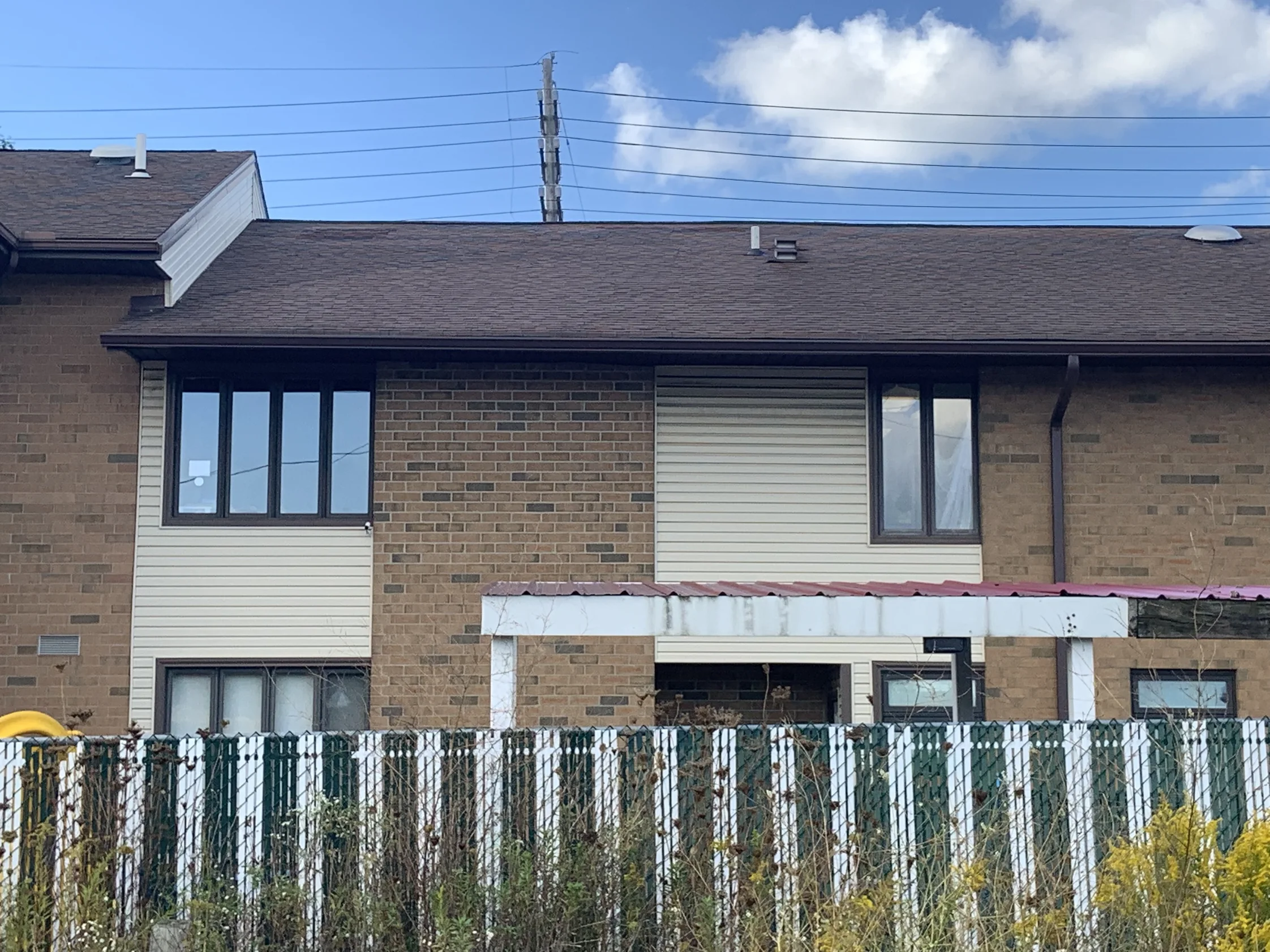 A long two-storey elevation in 2019, stained brick with darker units scattered through it between panels of cream siding, seen over a slatted boundary fence