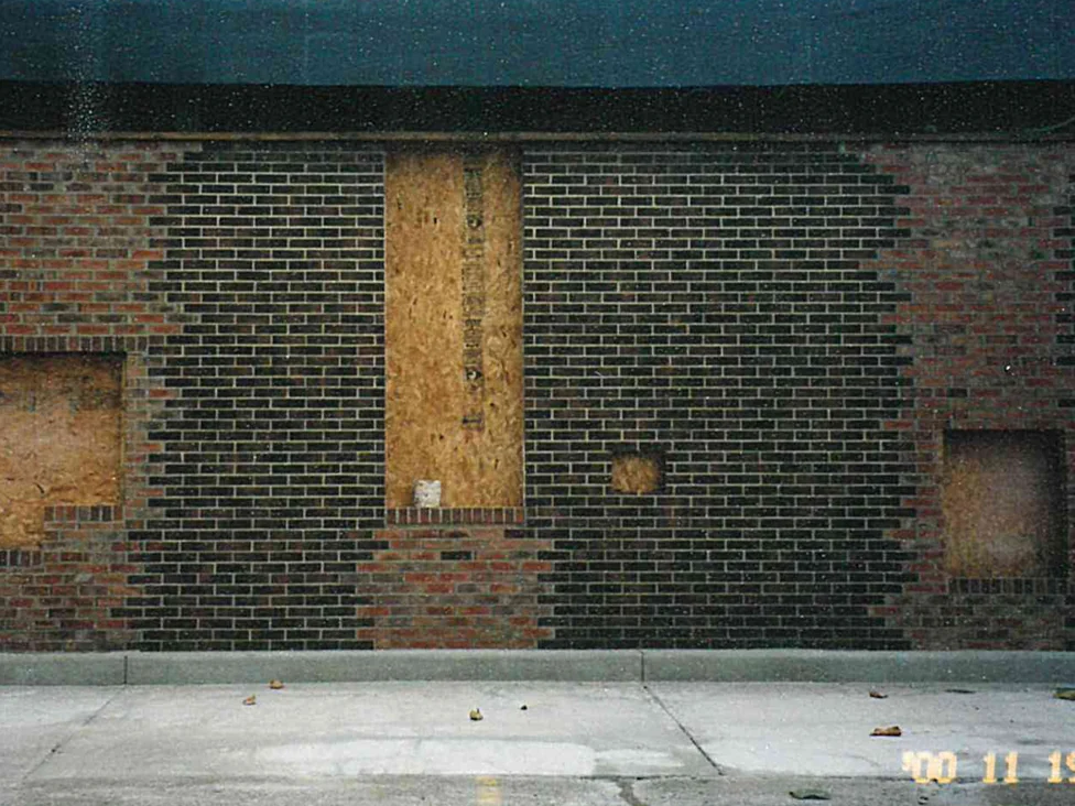 A long wall before staining, panels of pale red and buff brick set into a field of near black brick around boarded openings