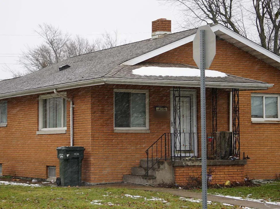 The front of a single-story orange brick house in 2020 with snow falling, the rebuilt corner beside the porch steps reading as the same brick as the rest of the wall