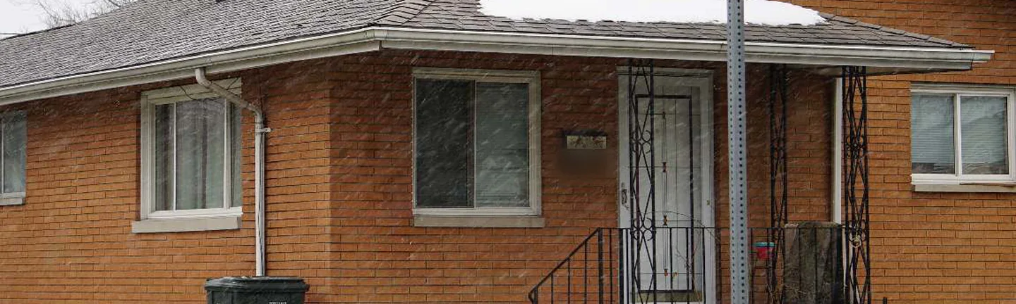 The front of a single-story orange brick house in 2020 with snow falling, the rebuilt corner beside the porch steps reading as the same brick as the rest of the wall