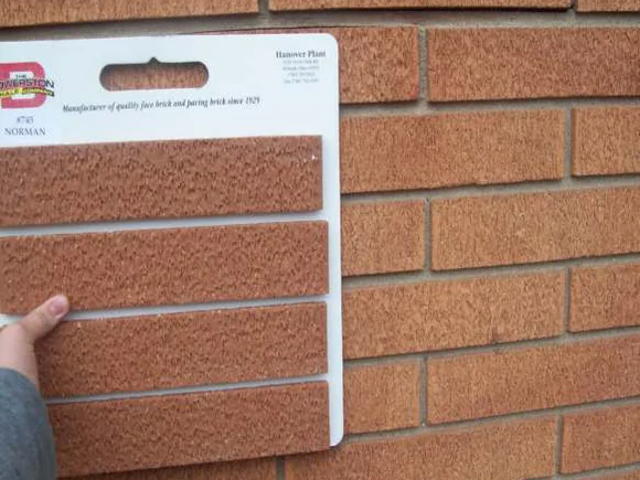 A hand holding a brick manufacturer's sample card of four replacement units against the original wall in 2010