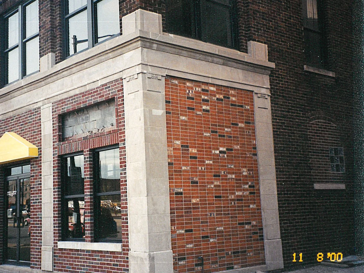 The corner of the building before staining, the alley wall peppered with pale buff and near-black units against the red brick of the street face