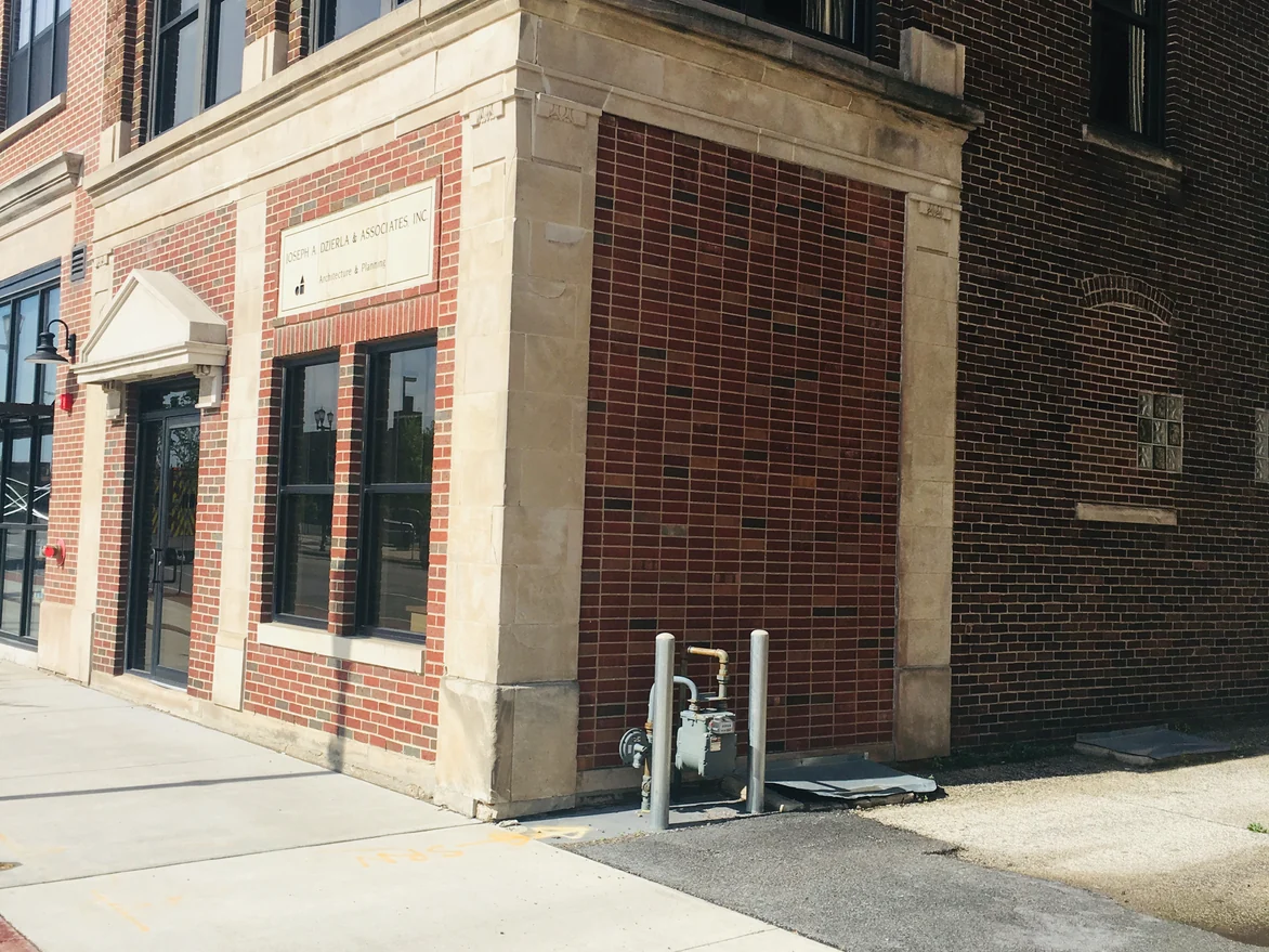 The corner seen from across the sidewalk in 2019, the stained alley wall and the red brick of the street face reading as one building