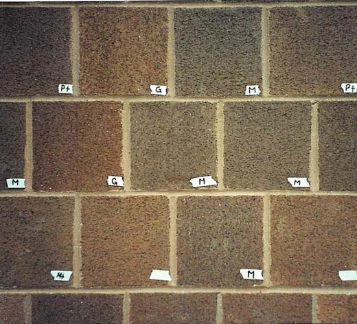 A brick test panel where individual bricks have each been stained a different shade, with small taped labels reading Pt, G, M and M plus under them