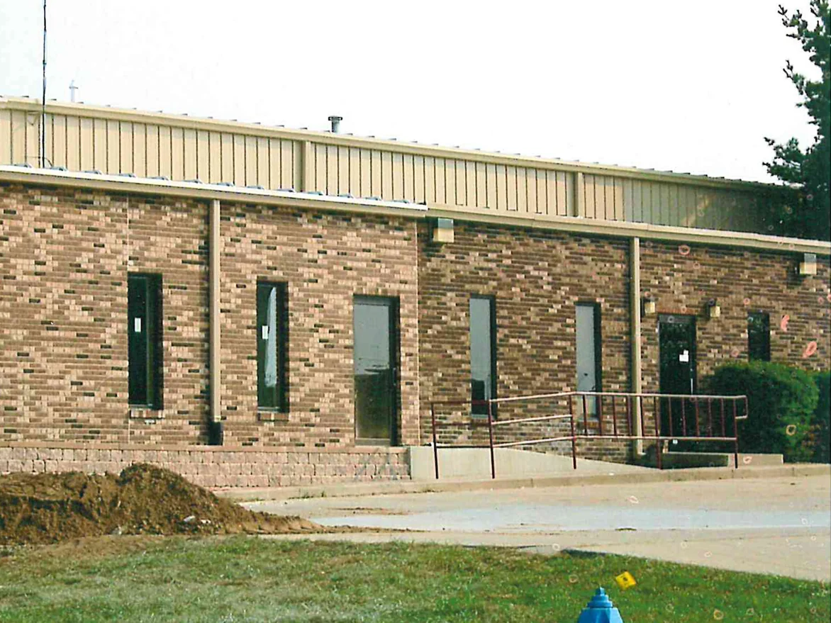 A single-story office in 2003, its brick a pale buff and tan mix under a beige metal upper wall, with fresh earth piled at the kerb