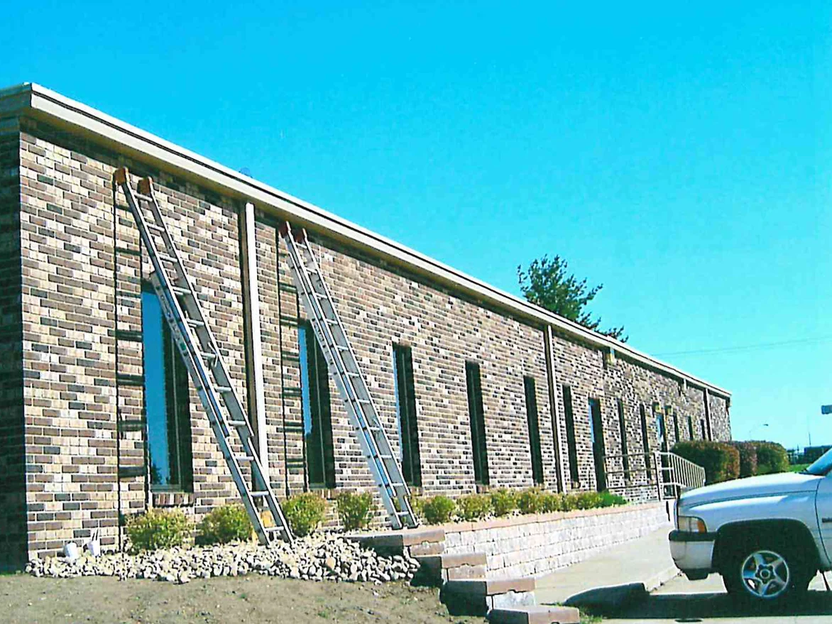 Two extension ladders standing against the same office during staining in 2003, the brick along the near bays still pale