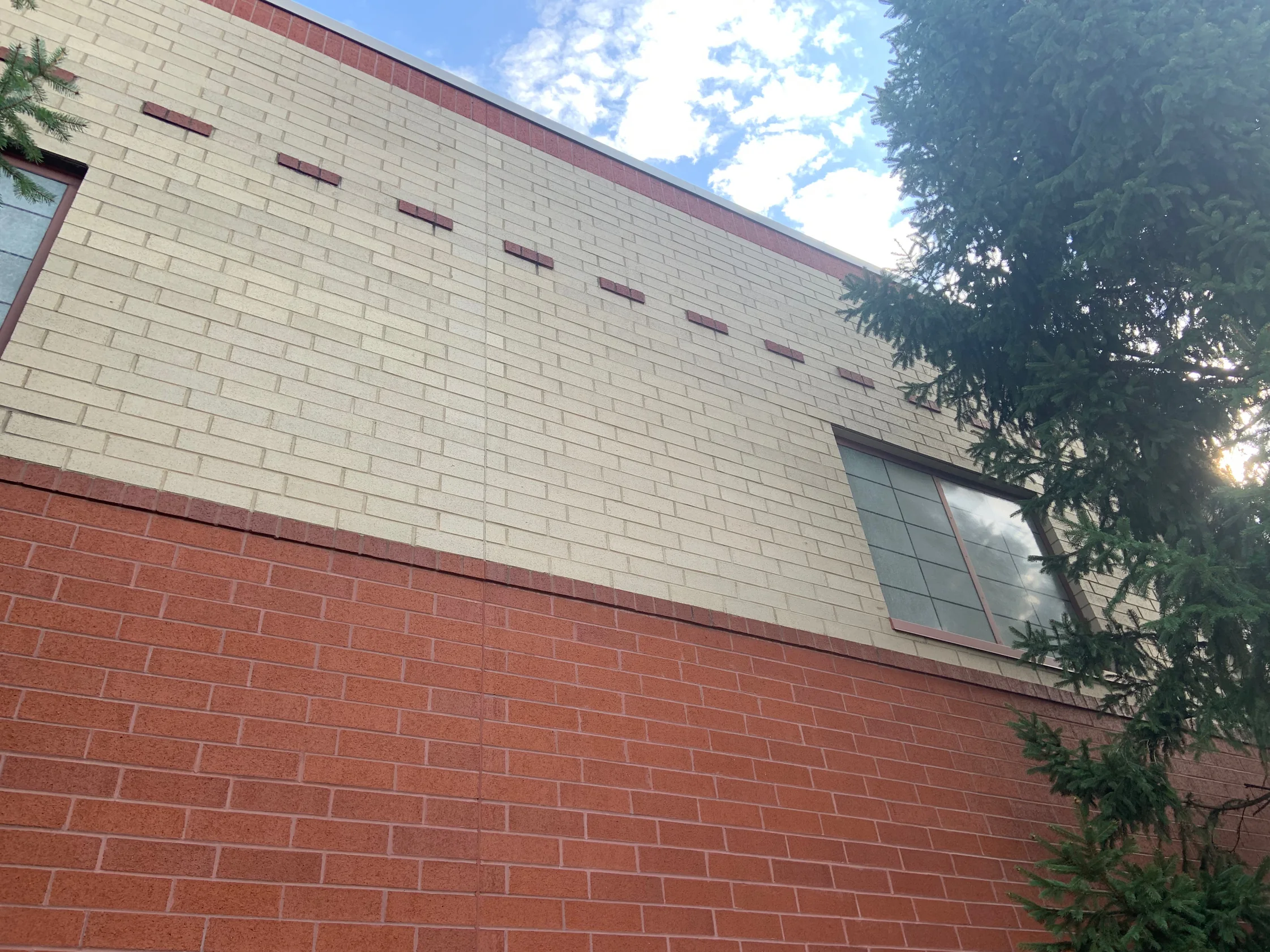 The school elevation in 2019, a cream brick field reading as one range above a red base band, with a dashed accent course and a window at the right