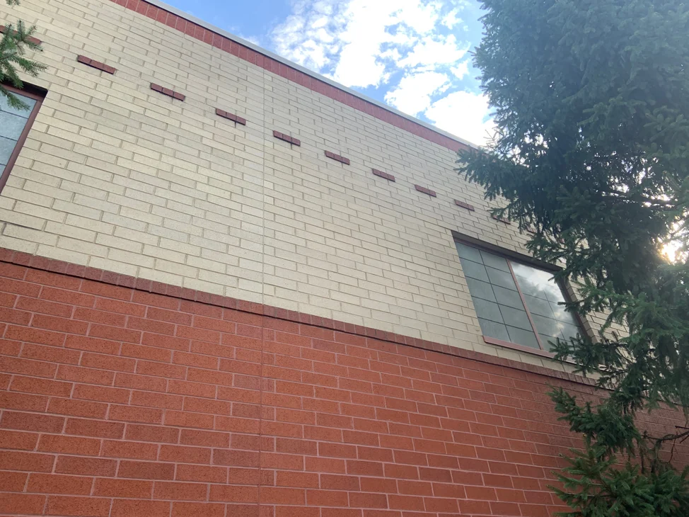 The school elevation in 2019, a cream brick field reading as one range above a red base band, with a dashed accent course and a window at the right.