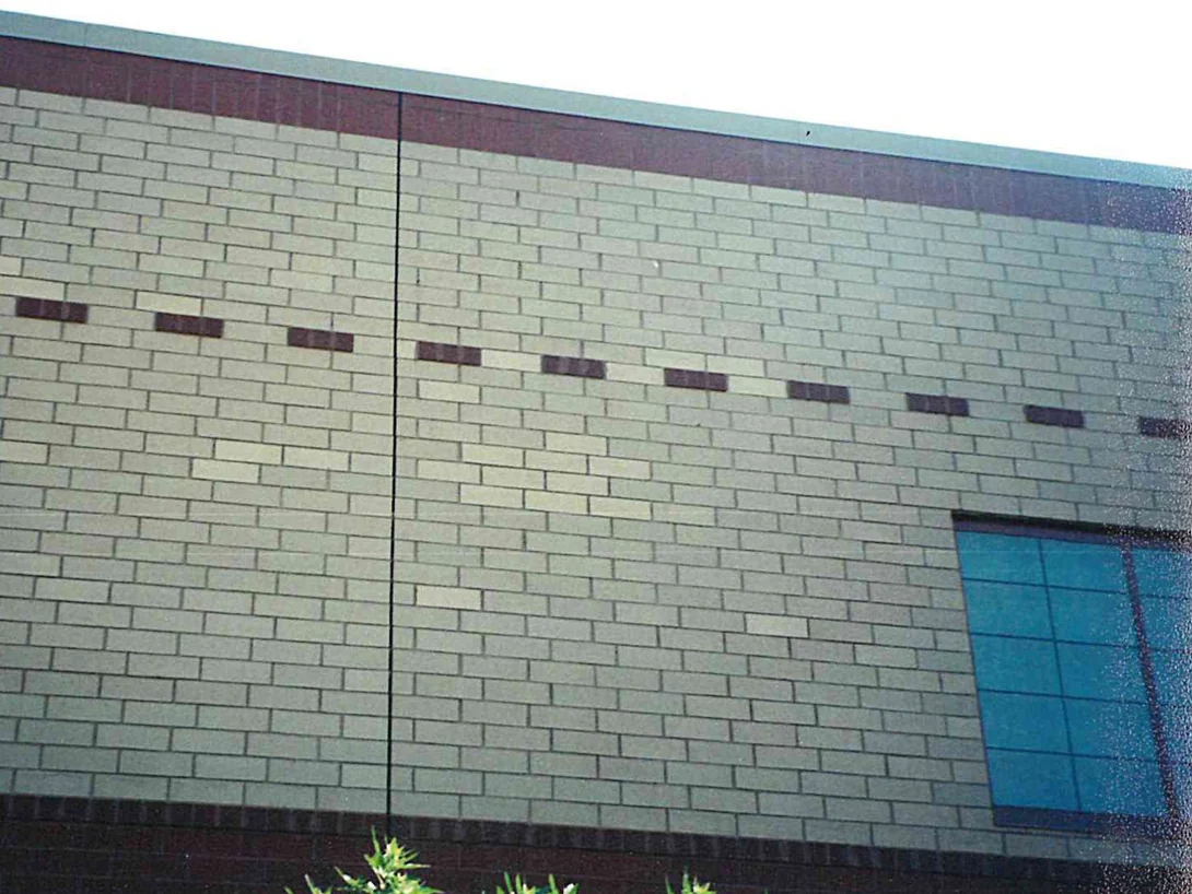 A pale cream brick wall above a dark base band, a dashed course of darker units running across it, with lighter bricks scattered through the field