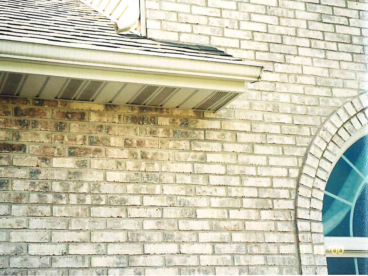 Close view of the wall under the eaves before the stain, a mottled brown and tan run of brick meeting a pale cream run beside an arched window