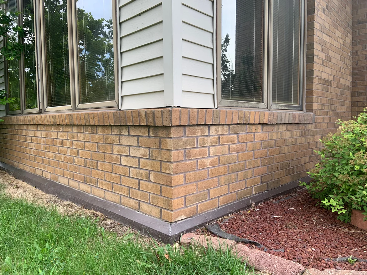 The outside corner of the low front wall in 2019, tan brick under a rowlock cap course, mottled darker across some units, with grass on one side and lava rock on the other