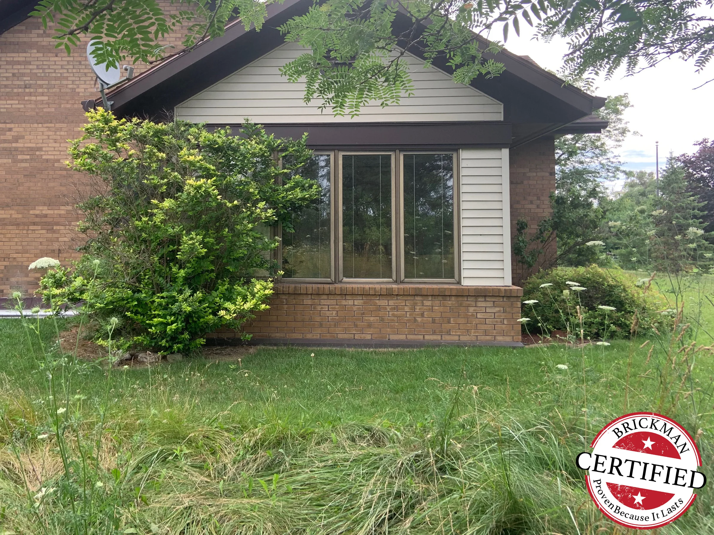 The gable end and glazed front bay of the house in 2019, the low wall under the windows reading the same brown range as the brick above it, with the Brickman certified stamp in the corner of the frame
