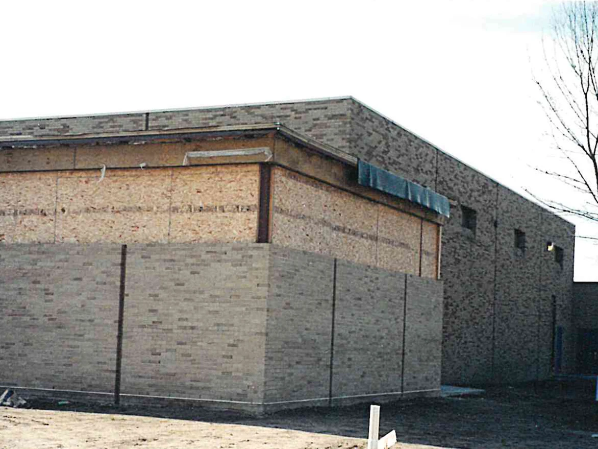 The corner of a new school wing in 2000, its brick an even light tan and cream, with oriented strand board sheathing still exposed above it and bare graded sand in front