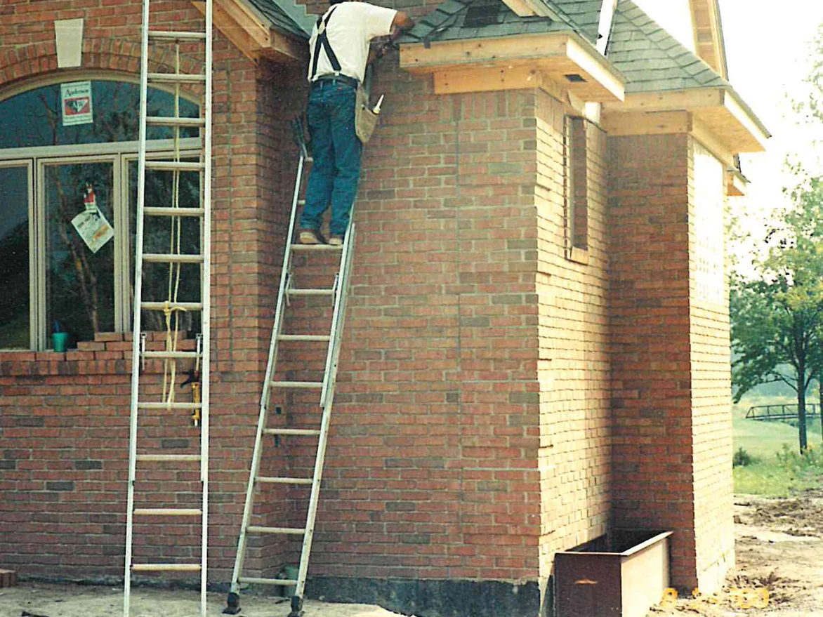 An outside corner of the house while it was still under construction, a worker up a ladder at the eaves, the front wall a deep red brown and the projecting wing beside it a much paler tan