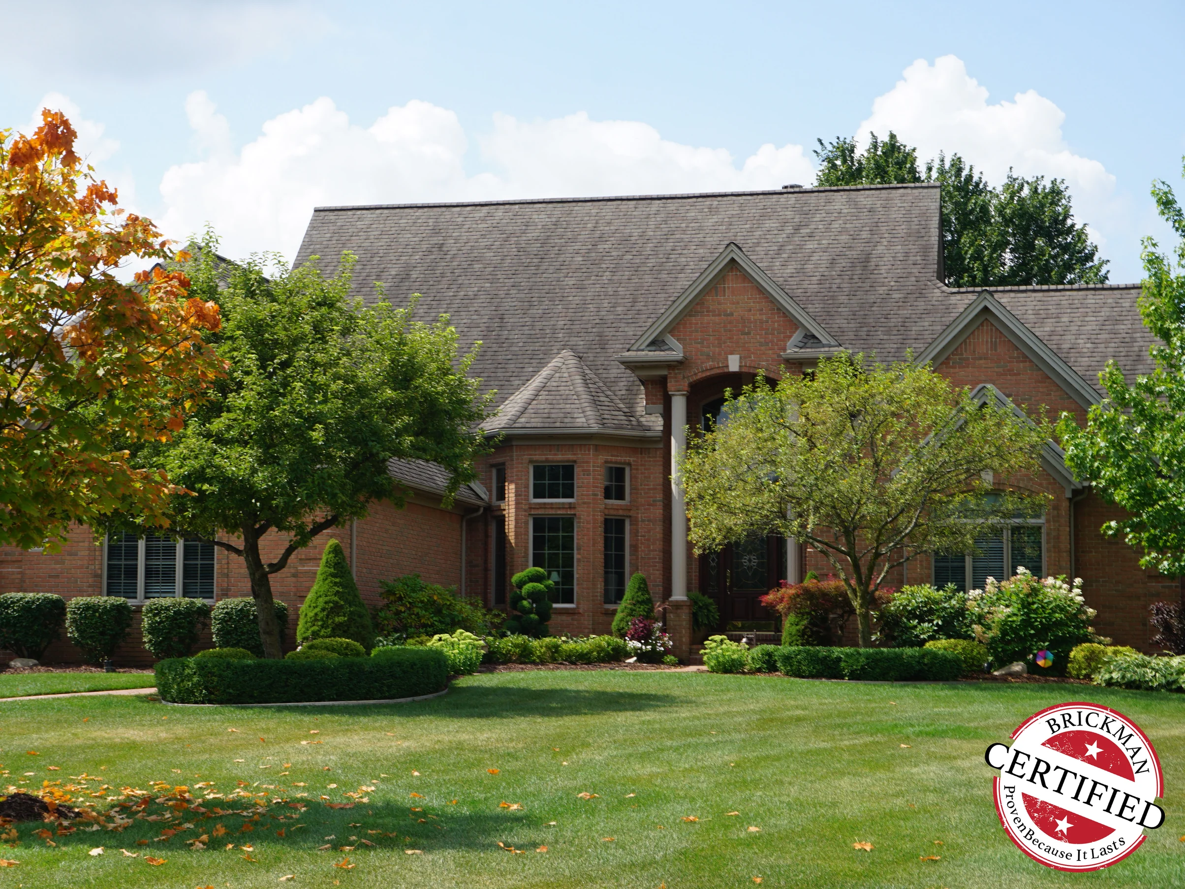 The house from the street in 2019 across a mown lawn, one even range of red brick running through the gables, the entry arch and the bay window, with the Brickman Certified stamp on the frame