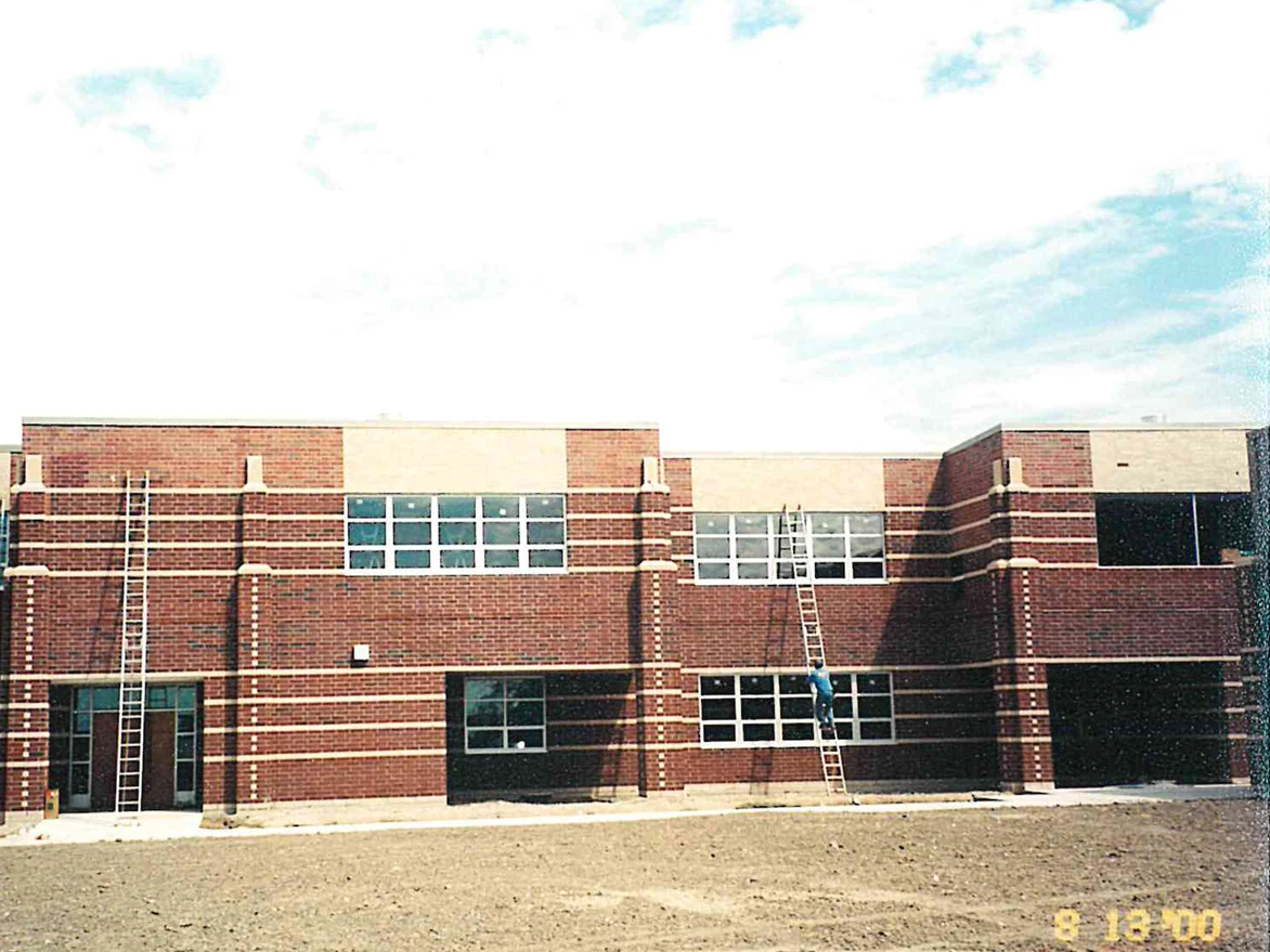 A two-storey school elevation in 2000, its red brick carrying paler orange and tan patches through the field, ladders leaning against the wall and bare graded ground in front