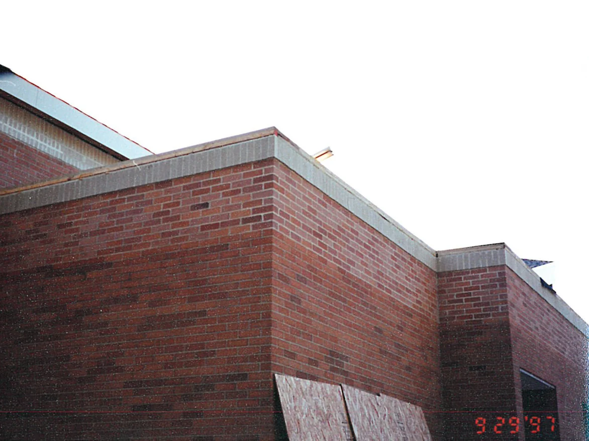 An outside corner of the church at parapet level in 1997, red and brown brick under a cream coping course, with board over an opening below