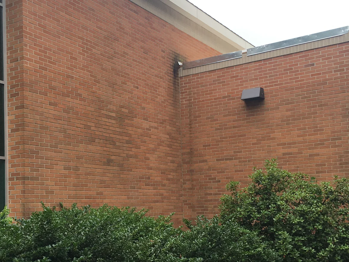 A tall brick wall meeting a lower wing in 2019, the brick weathered to one range with darker streaking under the roof edge