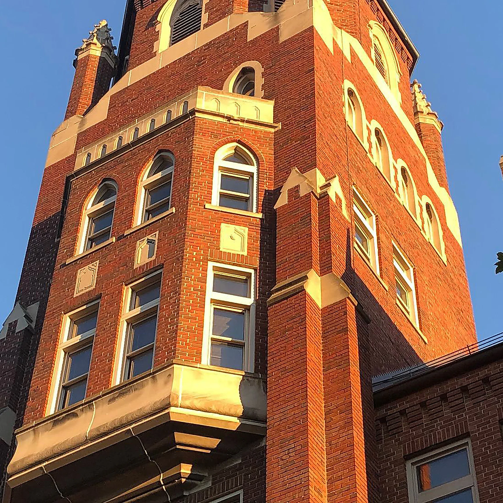The brick tower of the business school in 2020 in low evening sun, arched windows, limestone bands and a corbelled bay, the brick reading as one range from the pinnacles down to the roofline