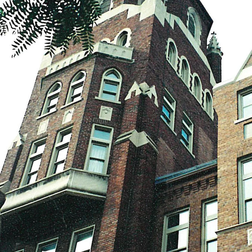 The brick tower of the business school in 2000, arched windows and limestone trim above a corbelled bay, shot from the ground through tree branches