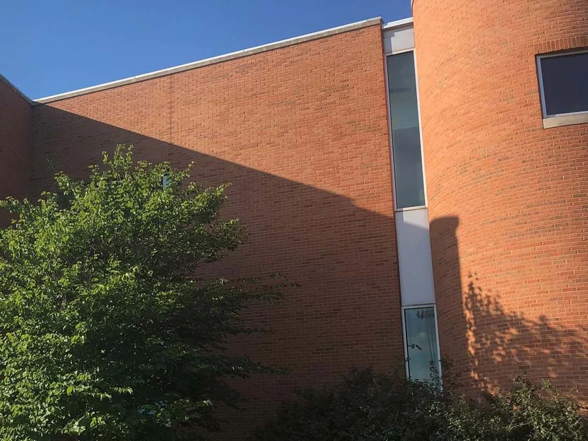 A curved corner of the building in 2019, orange brick running behind a tree past a strip window to the rounded end wall