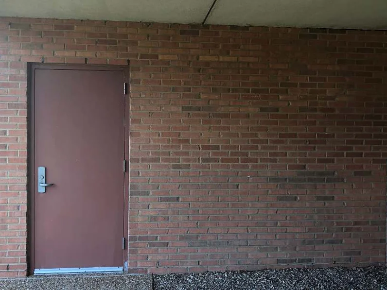 The same doorway in 2019, a maroon steel door set in a wall of even red brown brick under the soffit