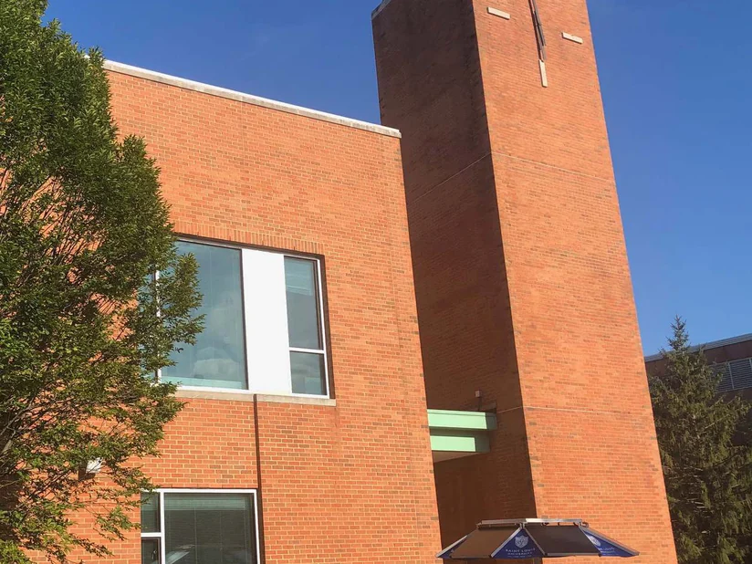 The tower and two-story wing of the university building in 2019, one range of orange red brick from the ground to the green glass pyramid, with a campus umbrella table at the entrance