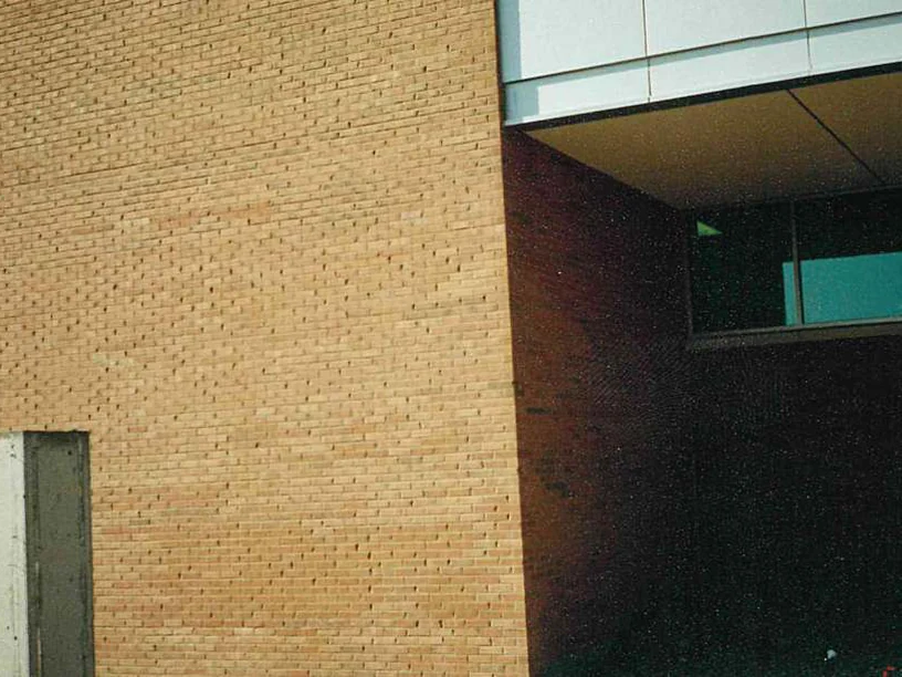 A pale tan brick wall in 1997 beside a dark recessed opening under a metal-clad soffit, with a strip window in the recess