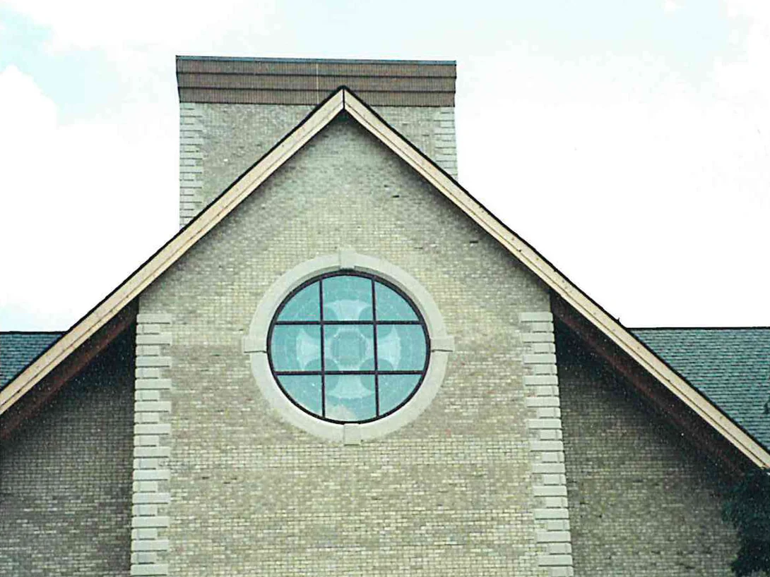 The gable end of the church with a round leaded window, its buff brick mottled with greyer off color areas