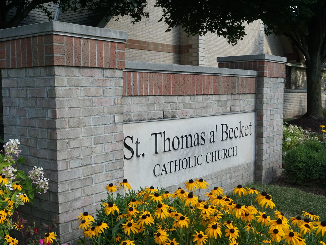 The brick monument sign at the entrance in 2019, lettered St. Thomas a' Becket Catholic Church, with black-eyed Susans in front of it