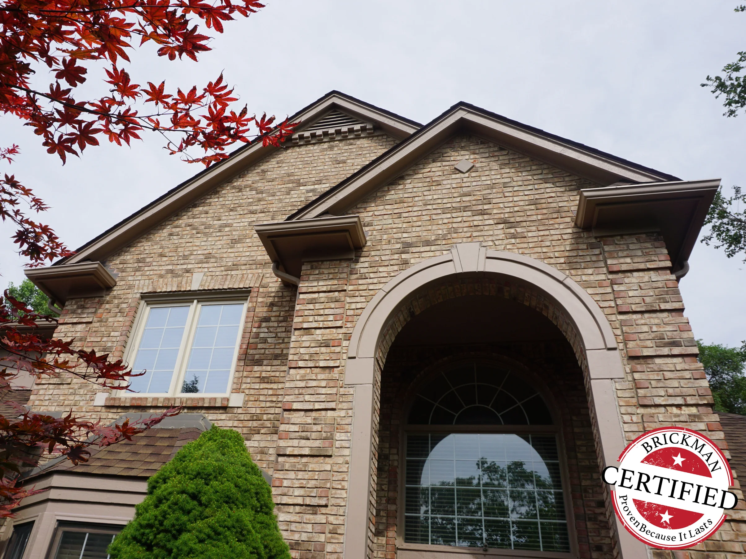 The gable and the arched entry seen from below in 2019, the brick an even tan and brown range, the Brickman certified stamp in the corner of the frame