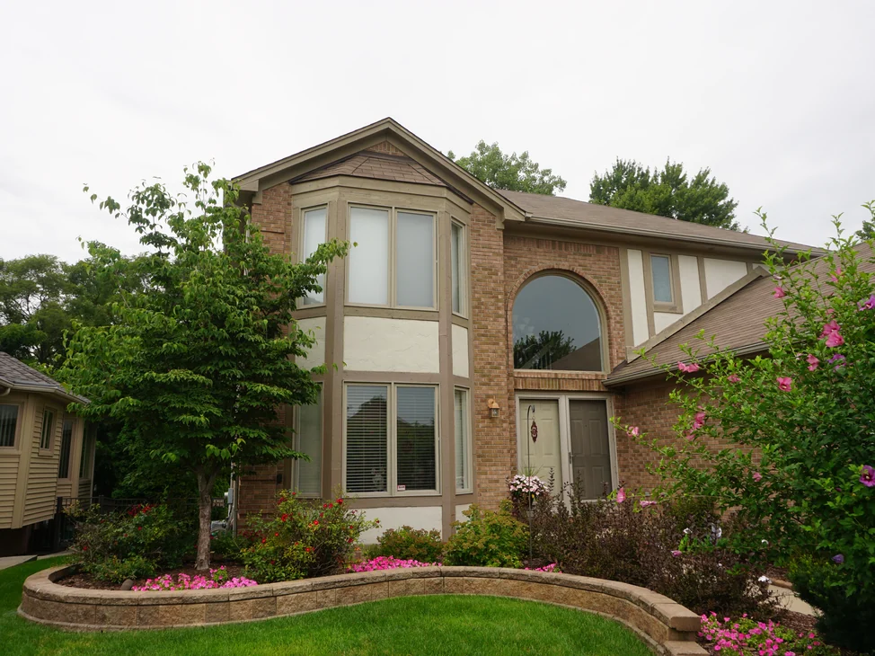 The house from the front lawn in 2019, the bay window, the arched window and the entry all carrying one warm tan brick blend