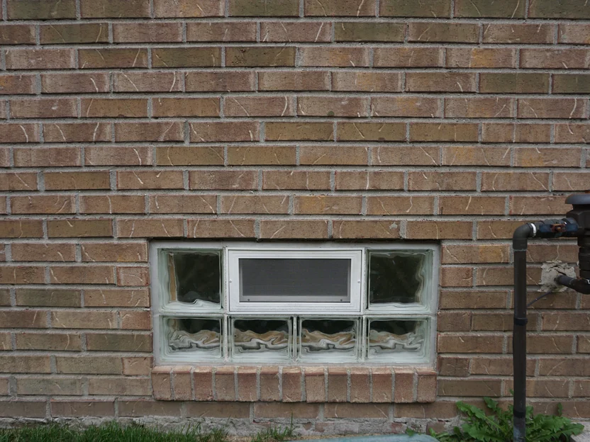 A basement window of glass block set in the wall in 2019, a rowlock sill beneath it and the brick even on both sides of the opening