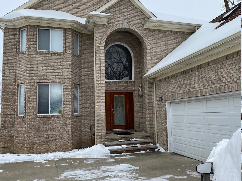 The front of the Sterling Heights house in 2021 under snow, tan brick running in one range across the bay, the arched entry and the attached garage