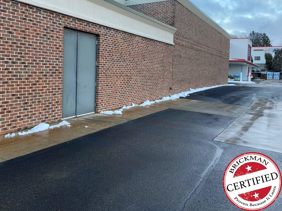The side wall of the store addition in winter 2020, red brown brick with a grey steel double door and snow along the base, with the certification stamp in the corner