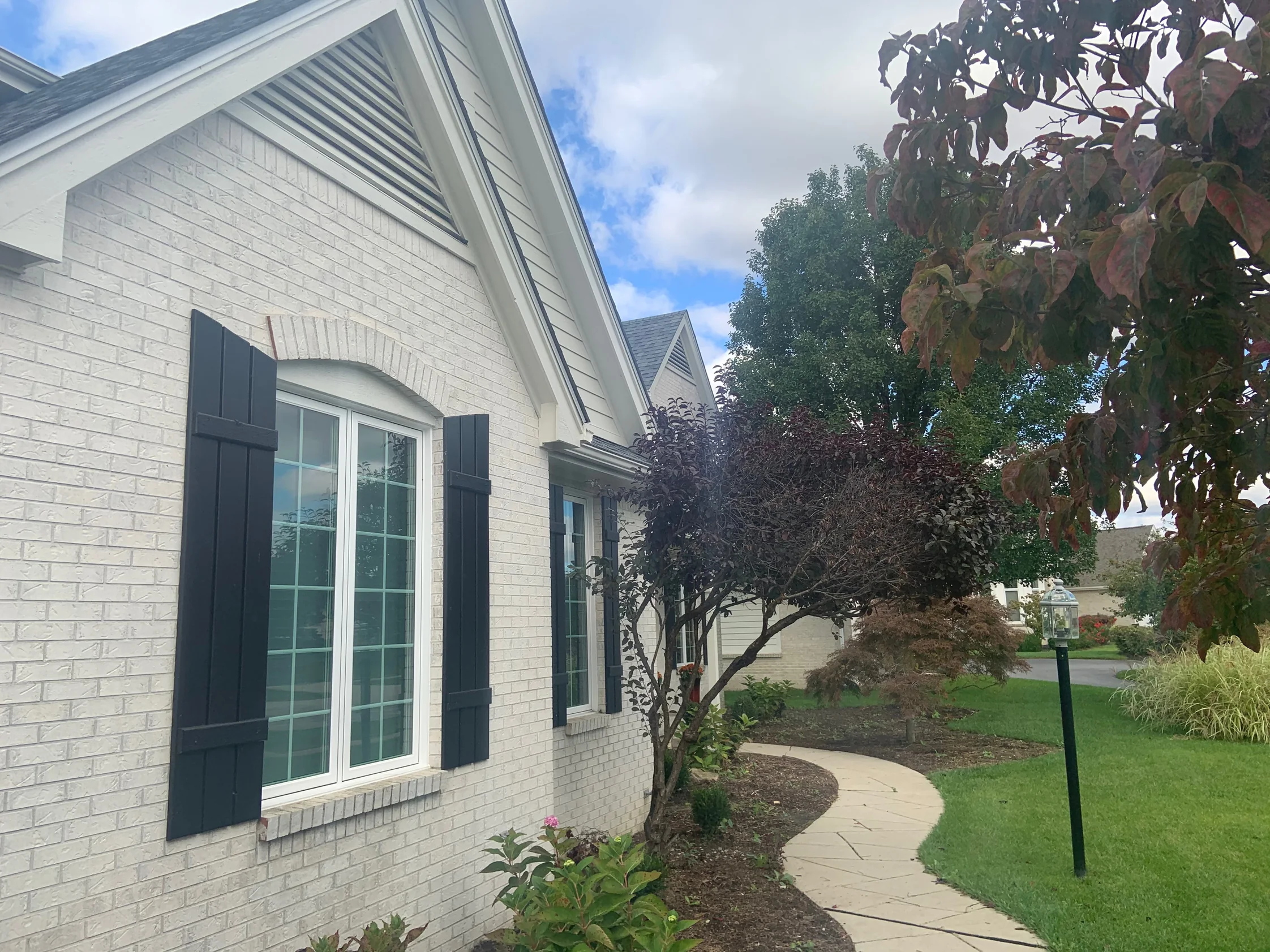 The side gable of the house in 2019, light brick around a shuttered window with a segmental brick arch, the front walk curving past it under a tree