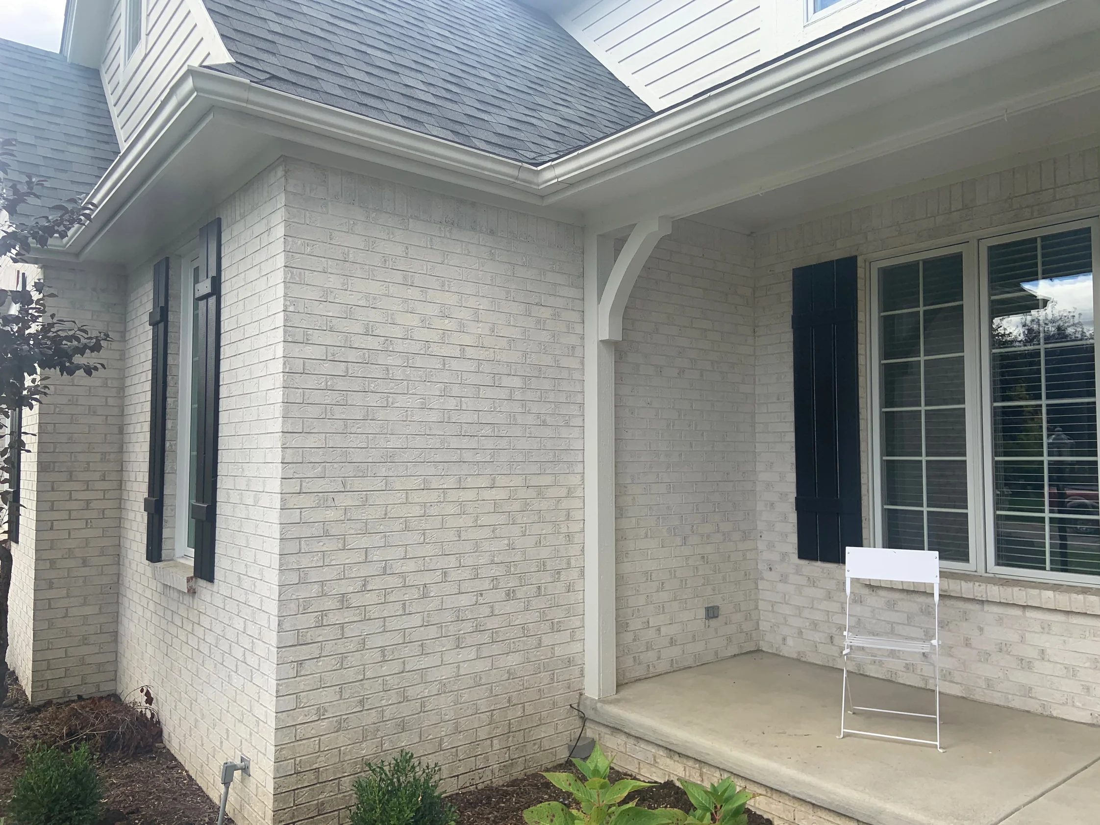 The porch corner in 2019, light brick running one range around the outside corner beside a white post and bracket, a black shutter on the left and the porch window on the right