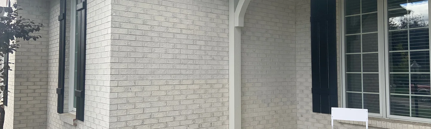The porch corner of the Sylvania house in 2019, light grey brick running one range around the corner past a white post and bracket and a black shutter
