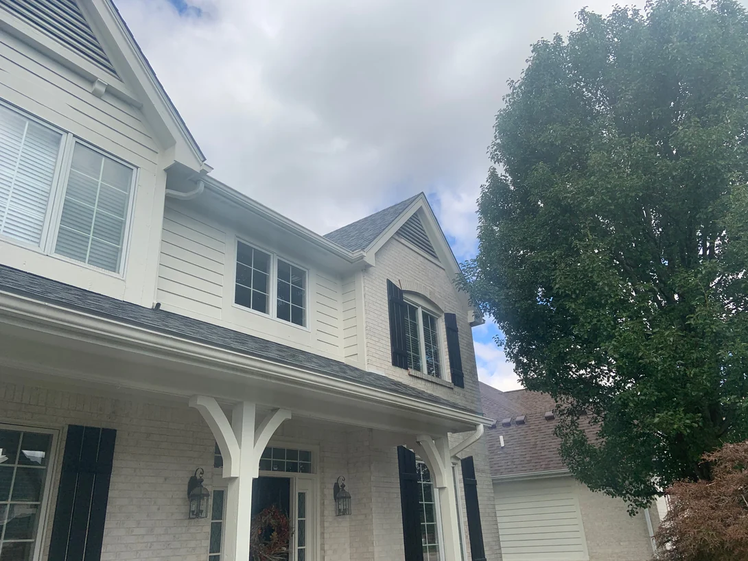 Looking up at the porch roof and the right-hand gable in 2019, light brick around the front door and the arched upstairs window under white siding