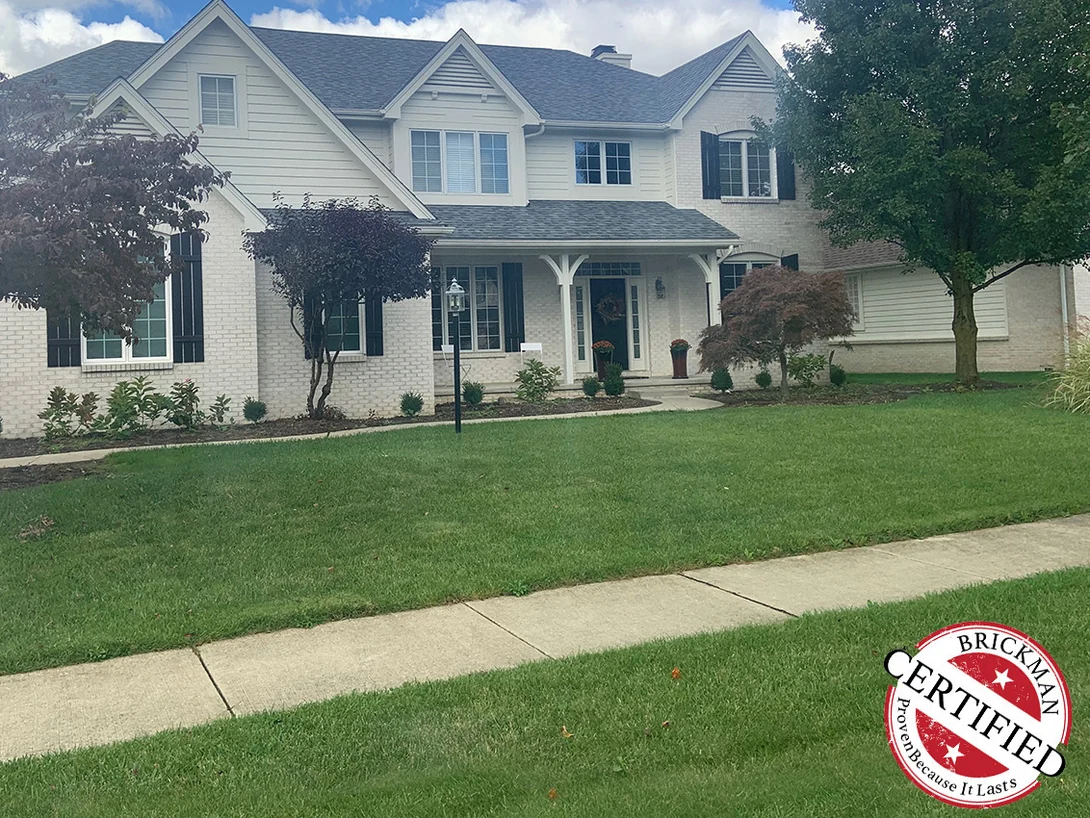 The whole front of the two-story house from the street in 2019, light brick under white siding gables and a grey roof, with the certification stamp in the corner