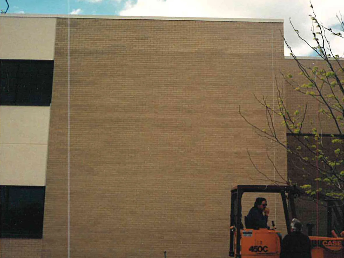 The blank buff brick end wall of the academy in 1999, with a tracked loader and two workers standing at the foot of it
