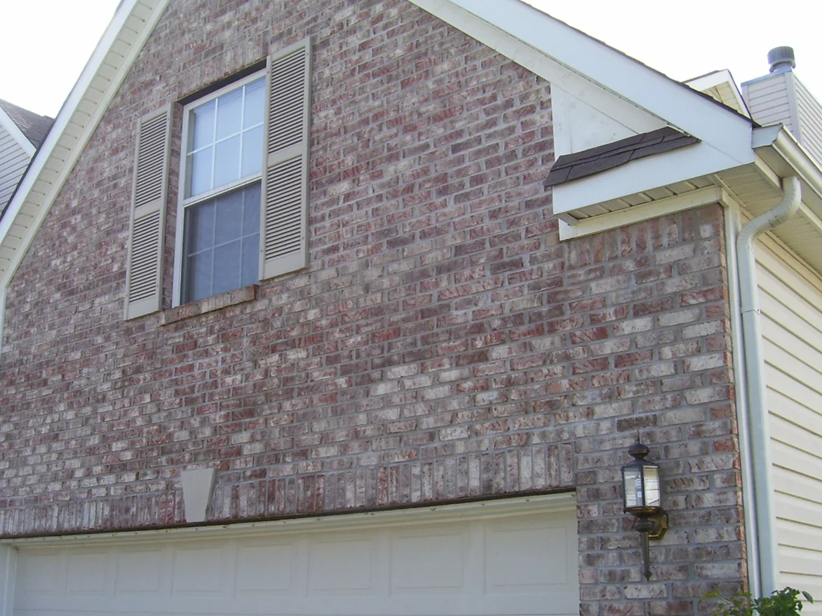 The garage gable in August 2009 before staining, its brick a run with pink and red through it under a shuttered window, a sliver of the main house's grey and white brick at the left edge